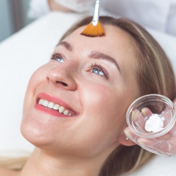 Mujer recibiendo un tratamiento facial en un spa, sonriendo mientras un especialista aplica una mascarilla en su frente con un pincel.