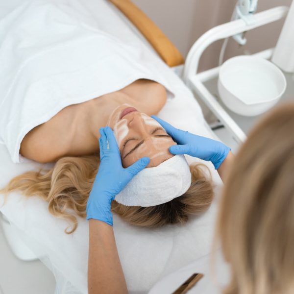 Mujer recibiendo un tratamiento facial en un spa, con una esteticista aplicando productos en su rostro mientras usa guantes azules.