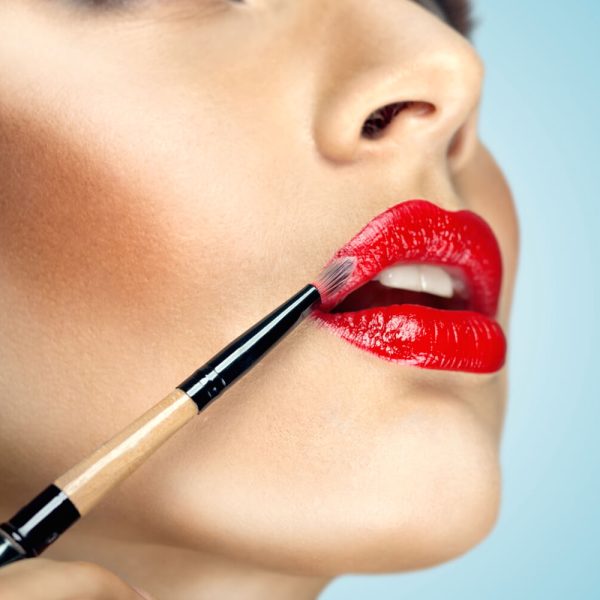 Mujer aplicando lápiz labial rojo brillante con un pincel, enfocado en su rostro y labios. Fondo suave que resalta el maquillaje.